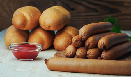 Ingredients for making homemade hot dogs. Sausages, fresh baked buns and ketchup sauce, served on wood chopping board over wooden background. Rustic styleの写真素材