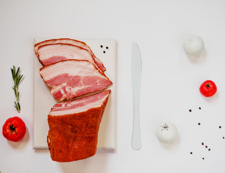 Smoked bacon on white cutting board with white colored tomatoes and knife on white backgroundの写真素材