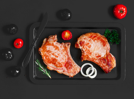 Baked pork steaks on a black baking sheet with black colored tomatoes on black backgroundの写真素材
