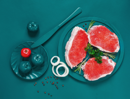 Raw fresh pork steaks on turquoise cutting board with turquoise colored tomatoes and knife on turquoise backgroundの写真素材
