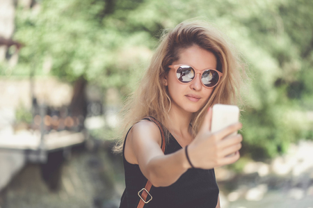 Closeup portrait of young happy smiling beautiful woman in sunglasses and black dress with long blonde curly hair doing selfie photosの写真素材