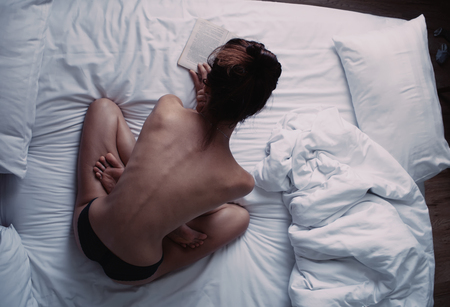 Young sexy naked girl reading book in bed in the morning, sitting in lotus position with her back to cameraの写真素材