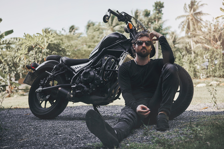 Handsome biker man in black wear sit near classic style cafe racer motorcycle. custom made motorcycleの写真素材