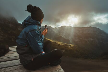 A hiker girl sits on the viewpoint and eats an energy bar, watching the sunset on the mountainsの写真素材
