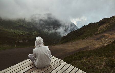 a girl in a grey hoodie sitting on a viewpoint in the mountainsの写真素材