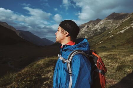 A young hiker in a blue jacket with orange backpack standing and looking up in the mountainsの写真素材