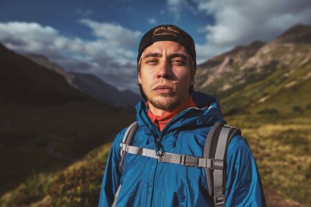 A young hiker in a blue jacket with backpack standing and looking in camera in the mountainsの写真素材