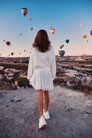 A tourist girl wearing white sweater on a mountain top enjoying wonderful view of balloons in Cappadocia. Happy Travel in Turkey conceptの写真素材