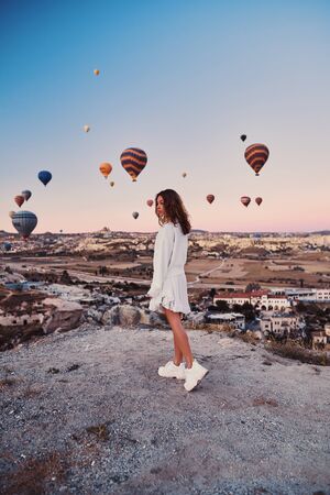 A tourist girl wearing white sweater on a mountain top enjoying wonderful view of balloons in Cappadocia. Happy Travel in Turkey conceptの写真素材