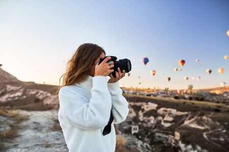 A tourist photographer girl wearing white sweater on a mountain top enjoying wonderful view and taking a pictures of balloons in Cappadocia. Travel photographer in Turkeyの写真素材