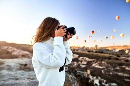 A tourist photographer girl wearing white sweater on a mountain top enjoying wonderful view and taking a pictures of balloons in Cappadocia. Travel photographer in Turkeyの写真素材