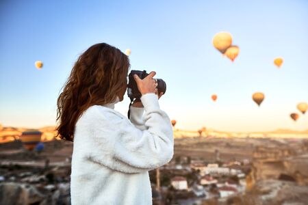 A tourist photographer girl wearing white sweater on a mountain top enjoying wonderful view and taking a pictures of balloons in Cappadocia. Travel photographer in Turkeyの写真素材