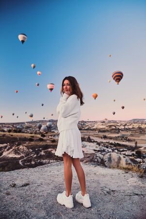 A tourist girl wearing white sweater on a mountain top enjoying wonderful view of balloons in Cappadociaの写真素材