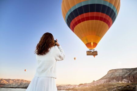 A tourist photographer girl wearing white sweater on a mountain top enjoying wonderful view and taking a picture of balloons in Cappadociaの写真素材