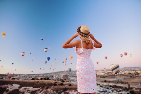 Young woman in dress and hat on a mountain top enjoying wonderful view of the sunrise and balloons in Cappadocia. Travel in Turkey conceptの写真素材