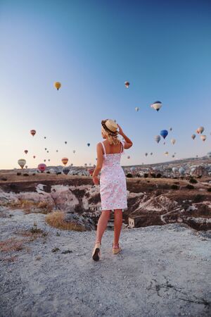 Young woman in dress and hat on a mountain top enjoying wonderful view of the sunrise and balloons in Cappadocia. Travel in Turkey conceptの写真素材