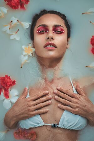 Fashion portrait of beautiful woman with red and white flowers in a bath with milk
