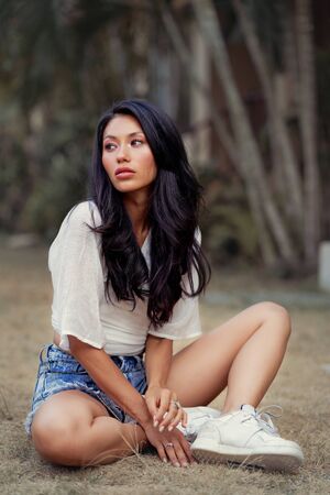 Beautiful Asian woman seats on grass, wearing jeans shorts, smiling enjoy weekendsの写真素材