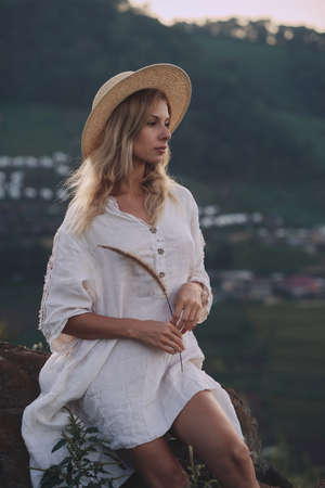 The blonde in a linen dress and straw hat sits on a viewpoint in the mountainsの写真素材
