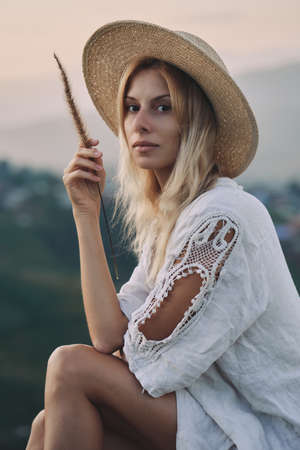 The blonde in a linen dress and straw hat sits on a viewpoint in the mountainsの写真素材