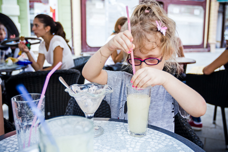 Little cute girl with glasses is drinking lemonade at pastry shop in summertime.の写真素材