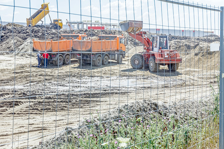 View through a fence wire on big loader until is filling a dump truck with soil at construction...