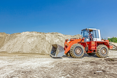 Red loader with wheels at earthmoving works, he is pushing sand on huge pile, preparing pile of for...