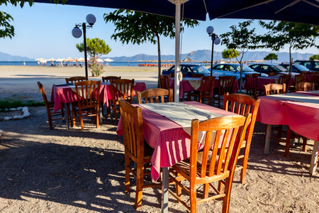 Chairs and tables in typical outdoor Greek tavern in morning sunlight with shadows on seashore.の写真素材