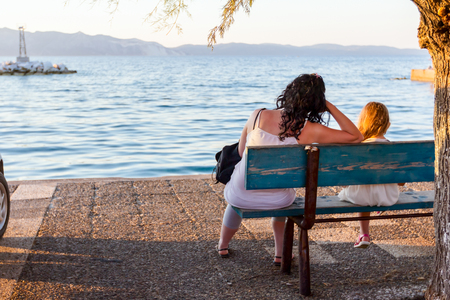 View of mother and daughter who are sitting on wooden bench facing to the open sea with islands in distance as silhouette across seashore.の写真素材