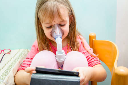 Child is take inhalation with mask on her face and play games with her phone.の写真素材