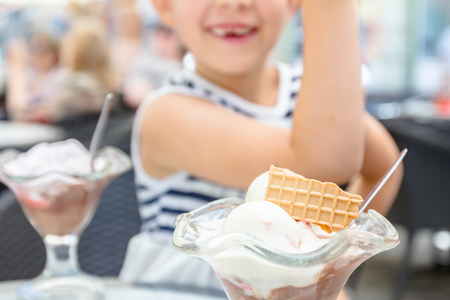 Little cute girl with glasses and carefree face expression is eating ice cream at table in pastry shop in summertime.の写真素材