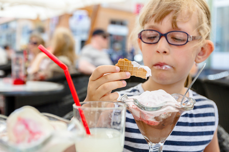 Little cute girl with glasses and carefree face expression is eating ice cream at table in pastry shop in summertime.の写真素材