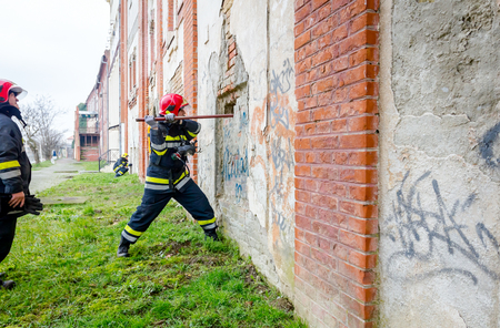 View from behind on firefighter in uniform with full safety gear, using crowbar to break brick wall.のeditorial素材