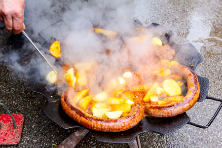 View through smoke on hand with metal tongs, turns the simmering potatoes and sausages on a barbecue grill.の写真素材