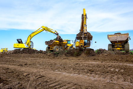 Big excavators are filling two dumper trucks with soil at construction site, project in progress.