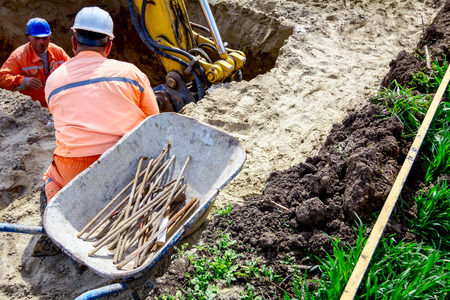 Group of workers with safety vests and helmets are help excavator to excavating square trench in...