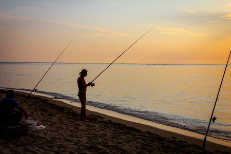 Two buddies are catching fish from the sandy beach with fishing rods, sunrise morning over Mediterranean sea.の写真素材