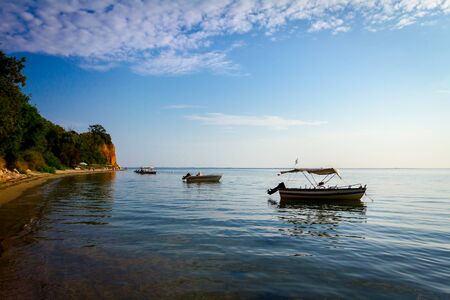 Anchored fisherman boats are on the water.  Cliff with vegetation is at sandy beach.