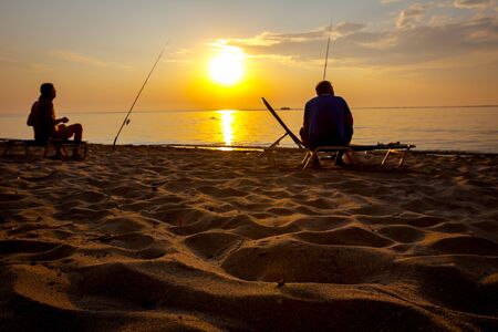 Two buddies are catching fish from the sandy beach with fishing rods, sunrise morning over...
