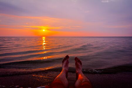 Man's legs until is sunbathing by lying carefree in lounger next to the coastline, on public beach.の写真素材