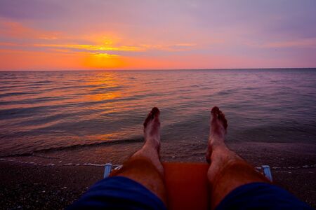 Man's legs until is sunbathing by lying carefree in lounger next to the coastline, on public beach.の写真素材