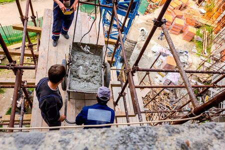 Workers are waiting crane to keep balance and direction to wheelbarrow, with mortar to placing on...