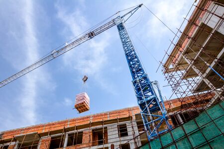 Crane is transport pack of red blocks over building site, raise up for building walls.