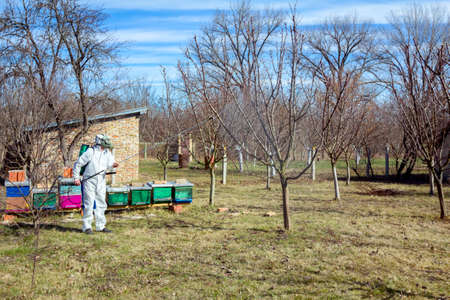 Farmer in protective clothing and gas mask sprays of fruit trees in orchard using long sprayer to...
