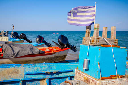 An old torn Greek flag flutters in the wind, set on a small wooden chair, fishing boats are dry...