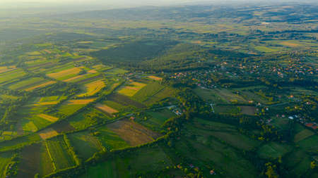 Shot above over green hilly landscape, several cultivated plots among forest trees and city in the...