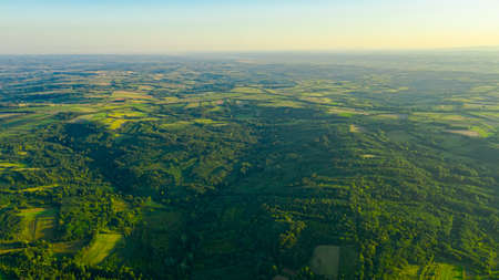 Above view over green hilly landscape, several cultivated, arable plots, among forest trees.