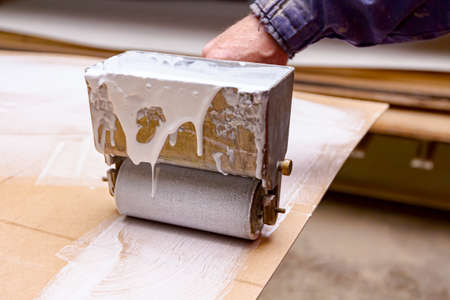 Close up shot on carpentry worker who uses handy roller tool, spreader for applying adhesive on the wooden surface profile, plywood that needs to be glued.の写真素材