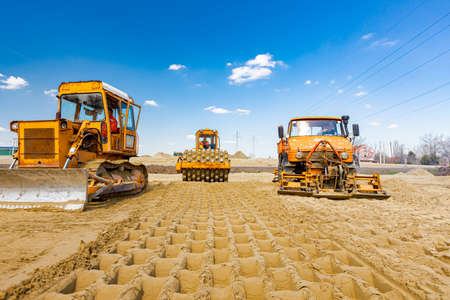 Road roller with spikes, bulldozer and truck with mounted plate vibration compactor are compacting,...
