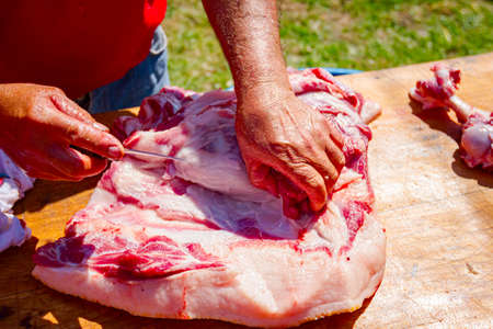 Butcher is cutting the raw pork meat into smaller pieces slice using sharp knife on a wooden board.